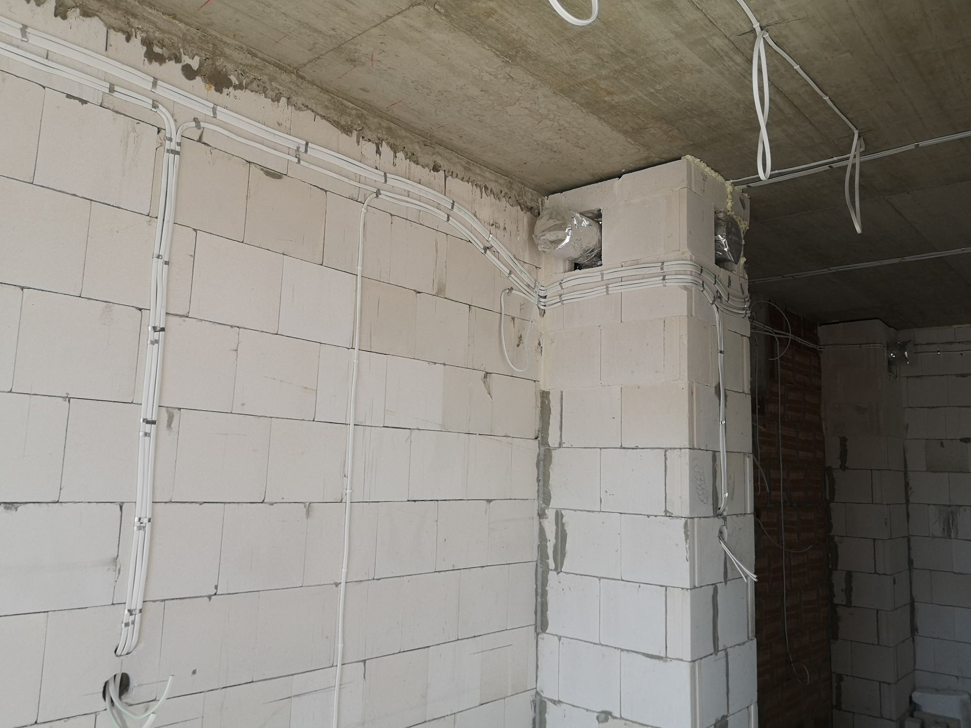 Białe przewody elektryczne prowadzone po ścianie z betonu komórkowego w surowym stanie, zabezpieczone metalowymi uchwytami, z widocznym otworem wentylacyjnym zabezpieczonym folią.