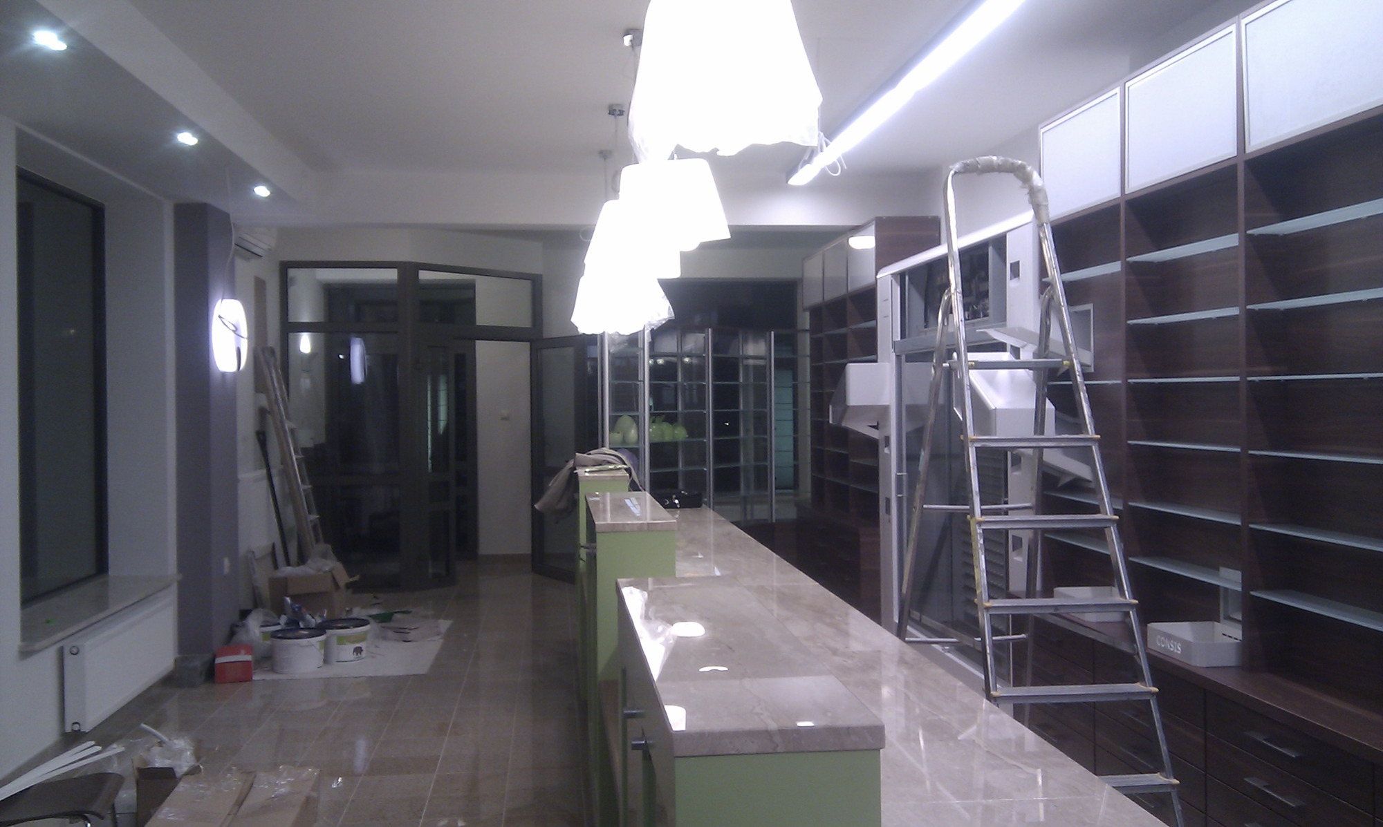 Wnętrze sklepu lub apteki w trakcie wykańczania: widoczne regały z ciemnego drewna, marmurowy blat lady, aluminiowa drabina, lampy pod sufitem i materiały budowlane na podłodze.