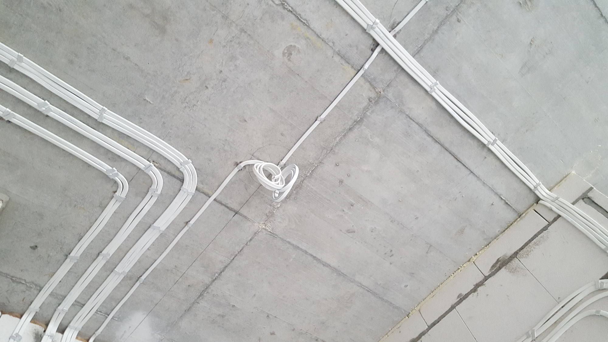 Białe rury instalacji elektrycznej przymocowane do surowego betonowego sufitu, widoczne kable zwinięte w spiralę.