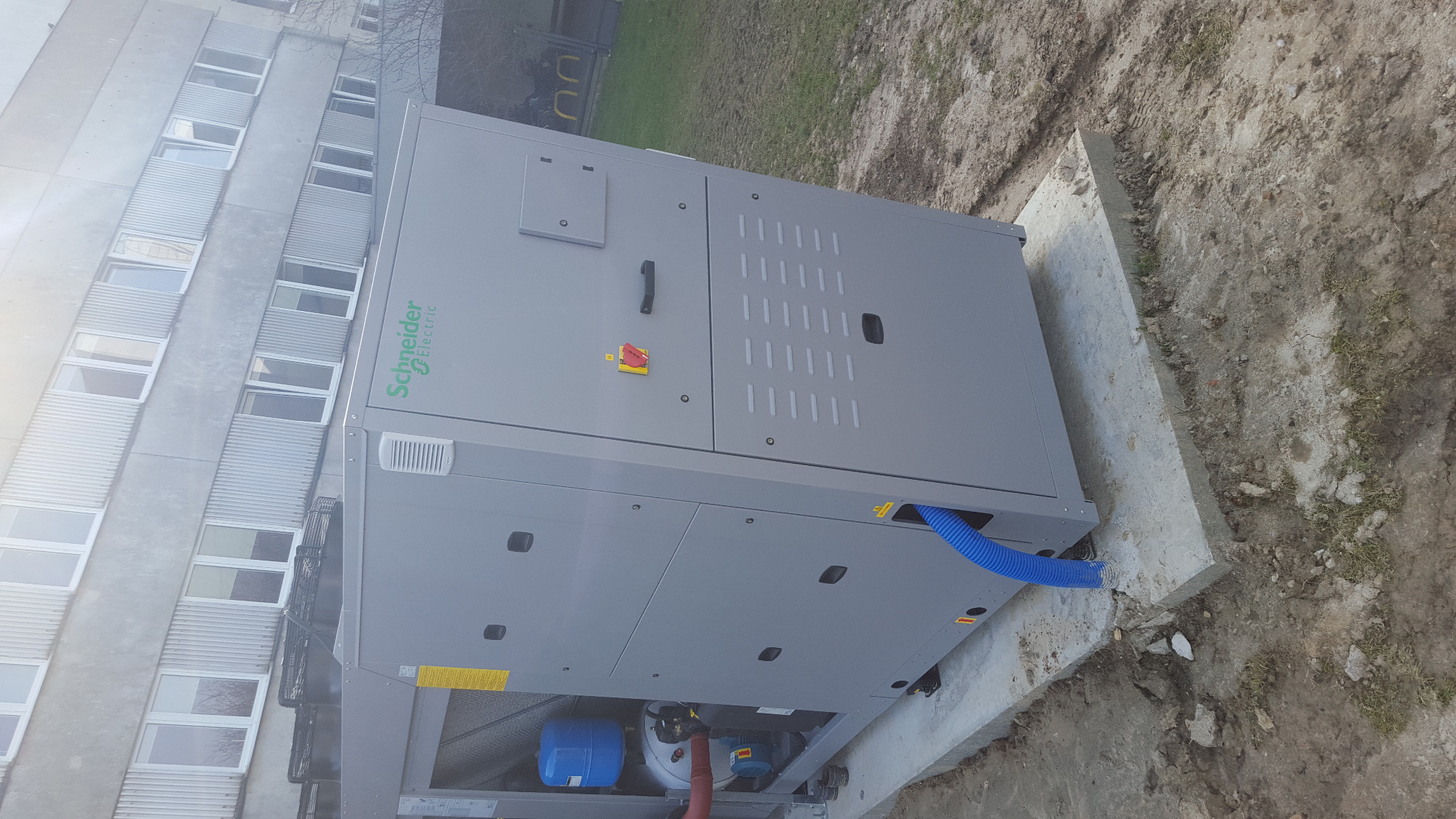 Szara, przemysłowa pompa ciepła Schneider Electric, zainstalowana na betonowej podstawie przy budynku, z widocznym niebieskim przewodem.