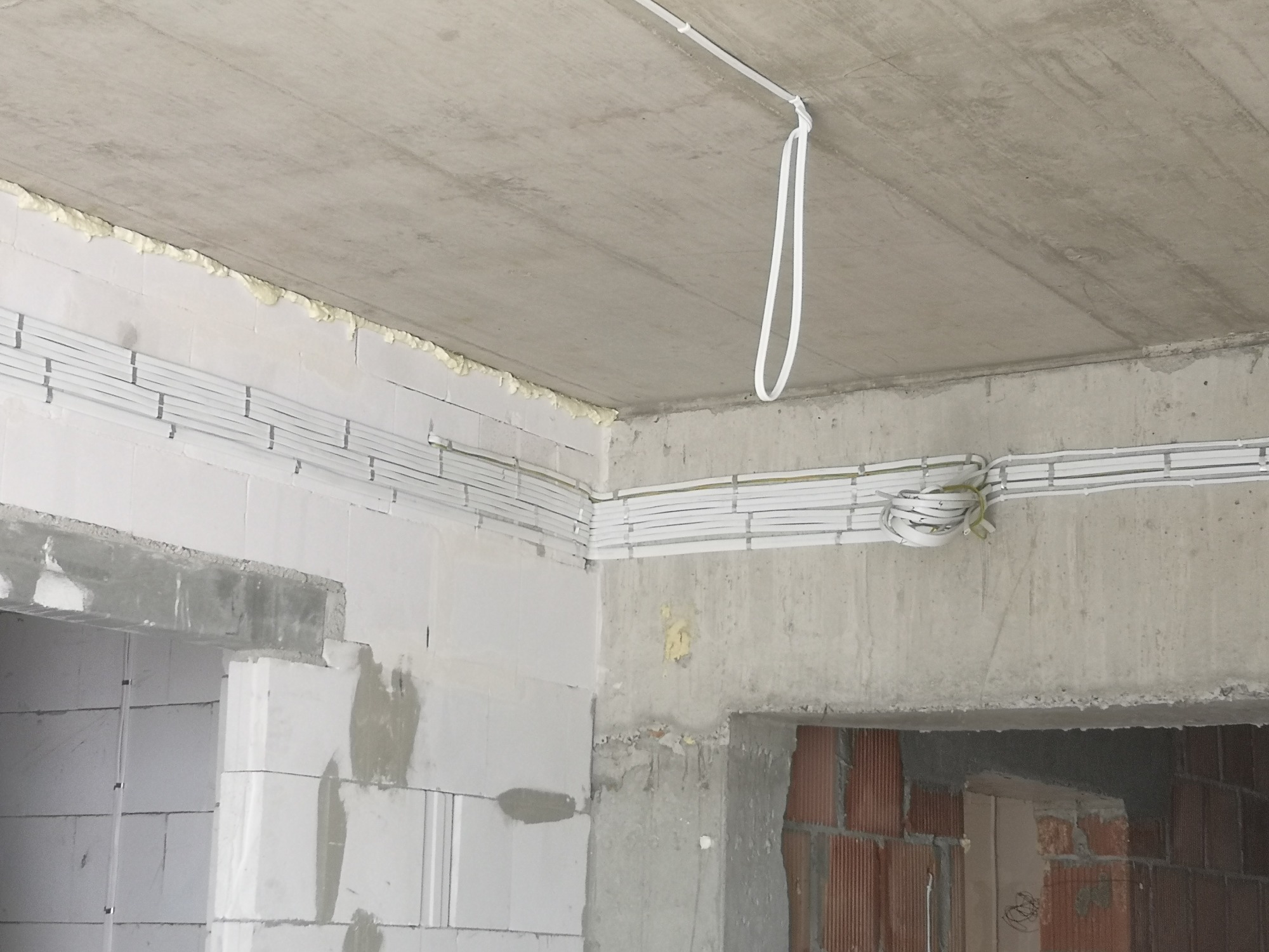 Wnętrze w stanie surowym z widocznymi białymi kablami elektrycznymi ułożonymi w korytkach na ścianach i suficie, z fragmentami pianki montażowej i surowymi otworami drzwiowymi.