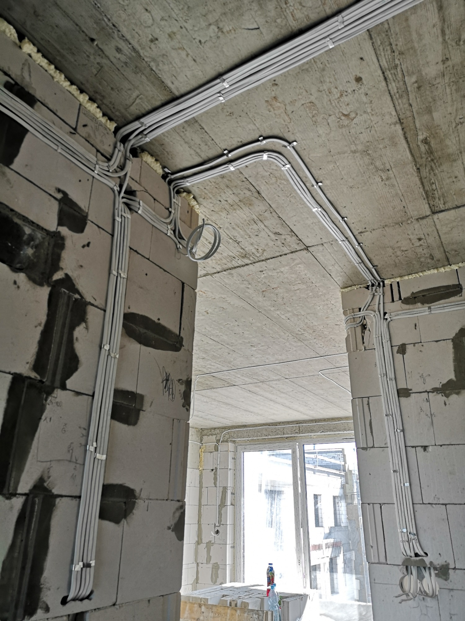 Szare kable elektryczne prowadzone po ścianach i suficie surowego wnętrza, zabezpieczone uchwytami, widoczne pianowanie szczelin przy stropie.