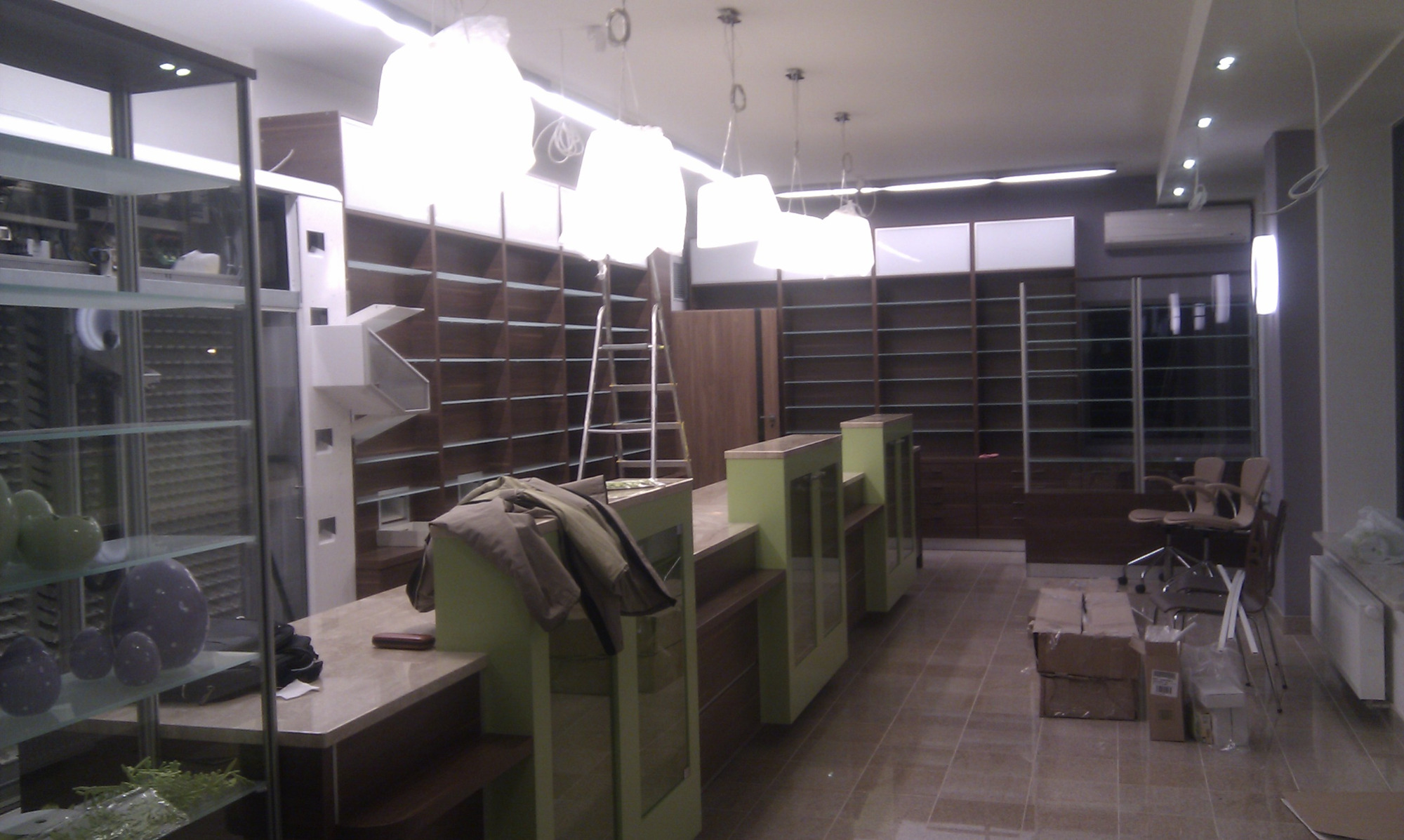 Wnętrze lokalu handlowego w trakcie aranżacji: regały z ciemnego drewna wzdłuż ścian, jasne lady sklepowe z zielonymi elementami, drabina oparta o regał, wiszące lampy w białych osłonach, kartony...