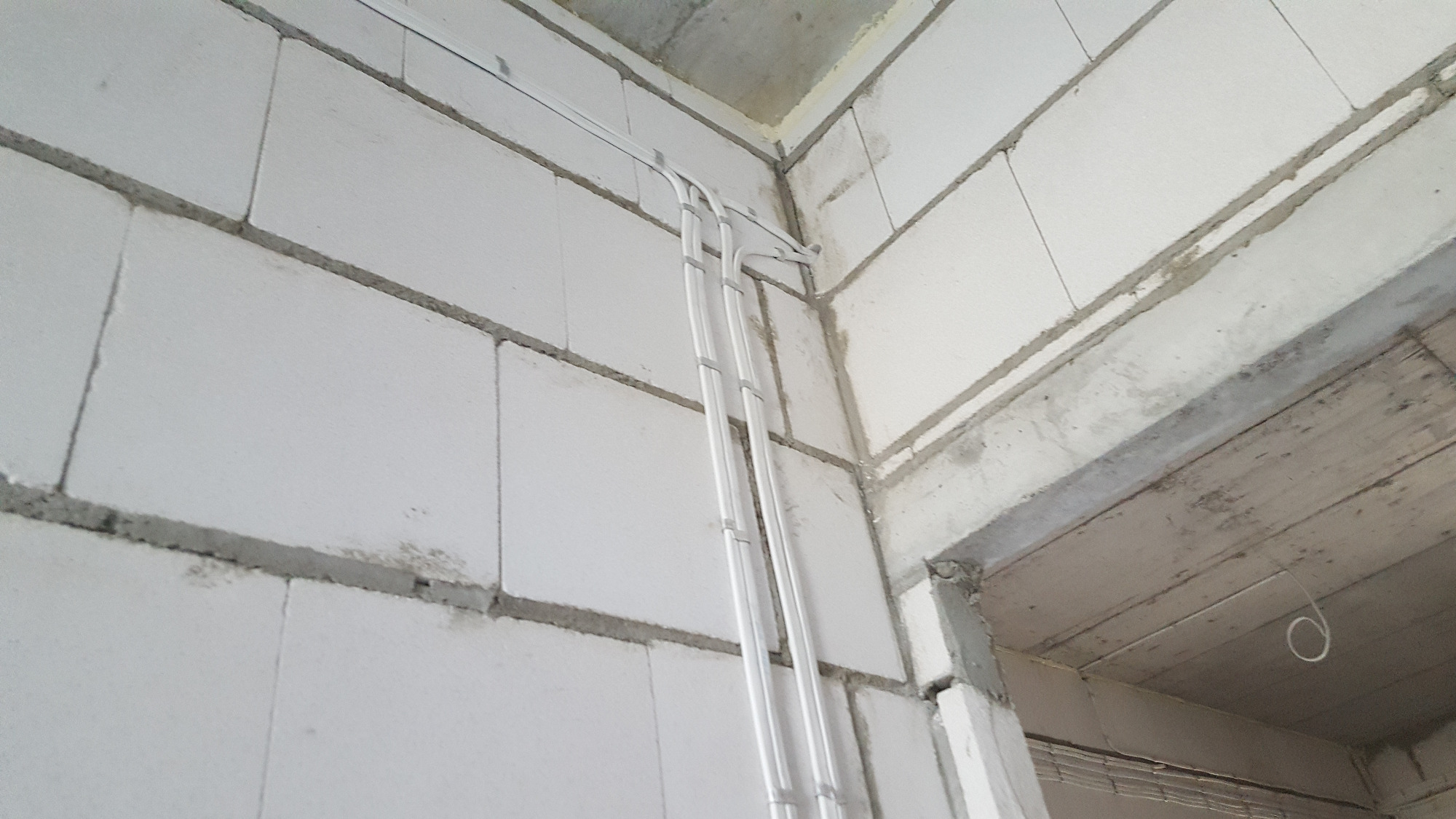 Białe rury instalacji elektrycznej prowadzone po ścianie z betonu komórkowego w surowym stanie, widoczne mocowania i zagięcia rur.