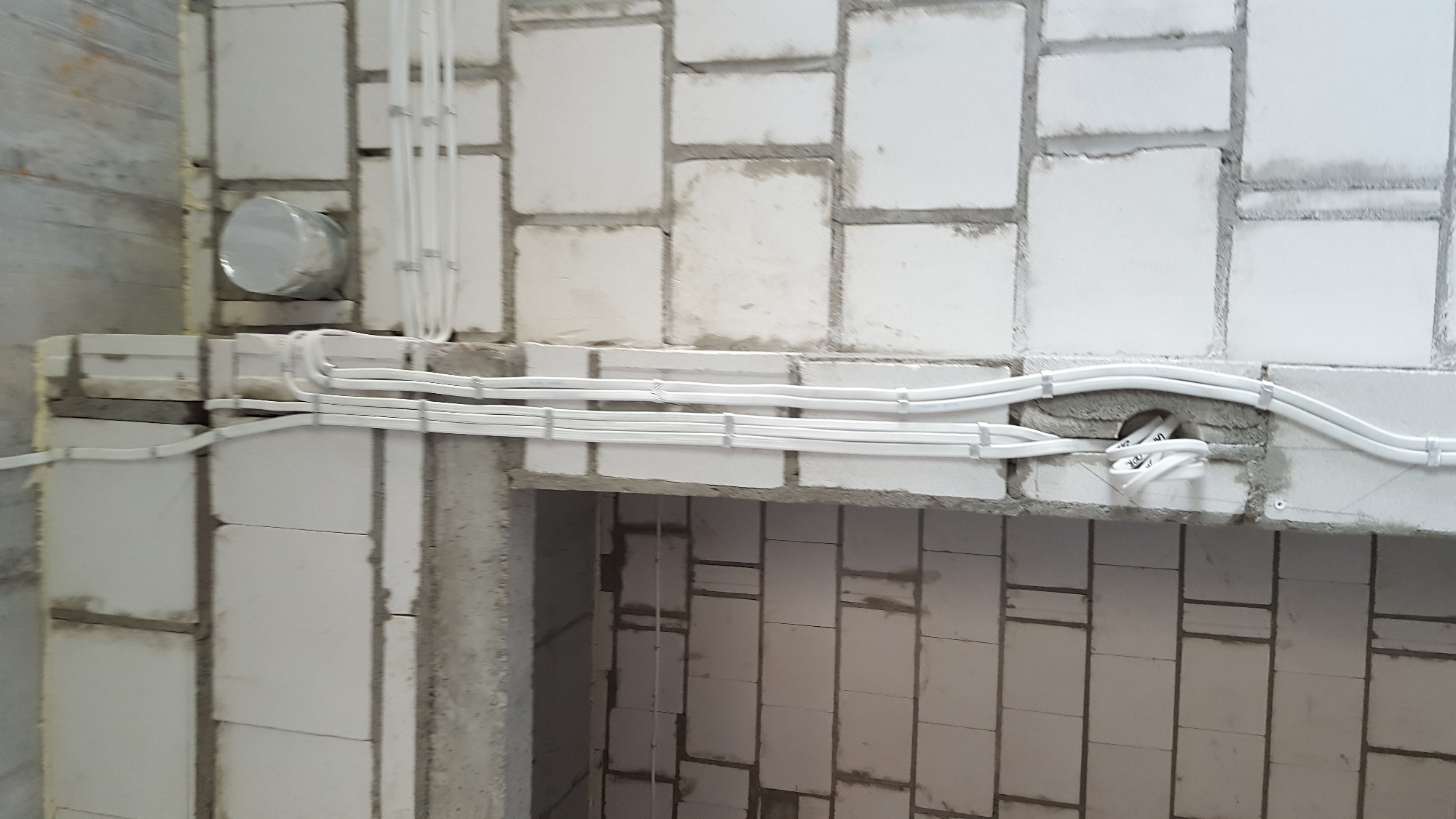 Białe kable elektryczne prowadzone wzdłuż ściany z białych bloczków, z widocznymi uchwytami mocującymi i puszką instalacyjną w trakcie montażu.