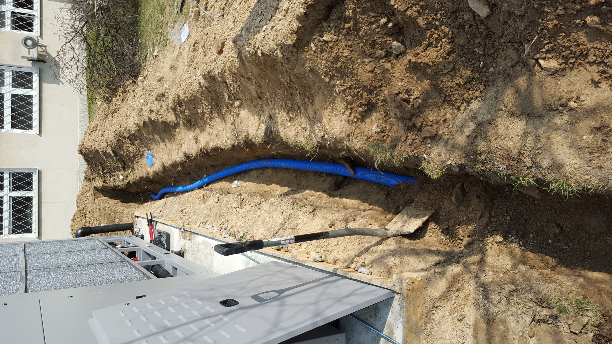 Niebieska rura karbowana ułożona w wykopie ziemnym, obok leży łopata z czarną rączką, widoczny fragment budynku z białymi oknami i klimatyzatorem na ścianie.