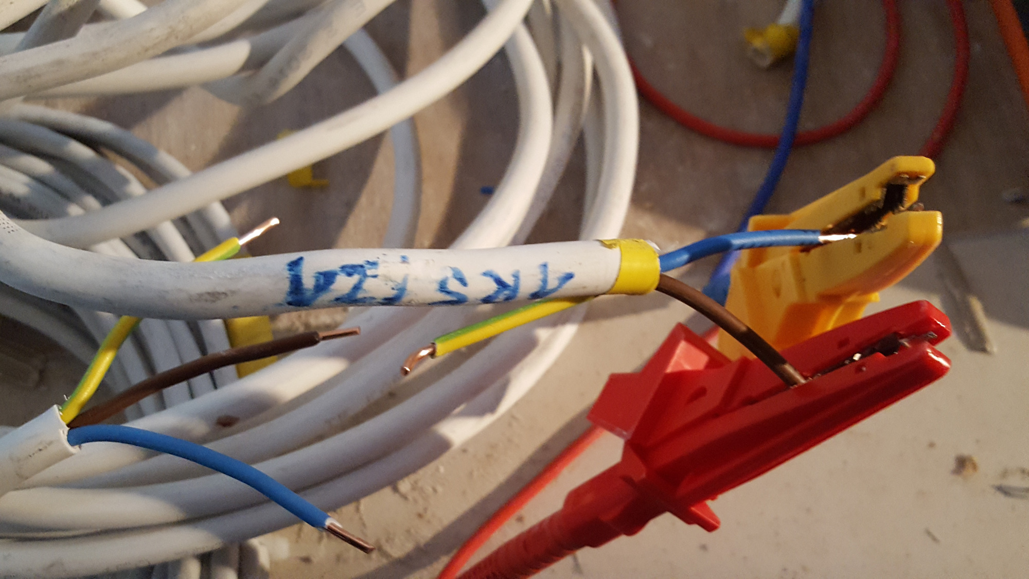 Fragment instalacji elektrycznej z przewodami o różnych kolorach, połączonymi za pomocą klem, na białym kablu widoczny niebieski napis '1KS FZ1'.