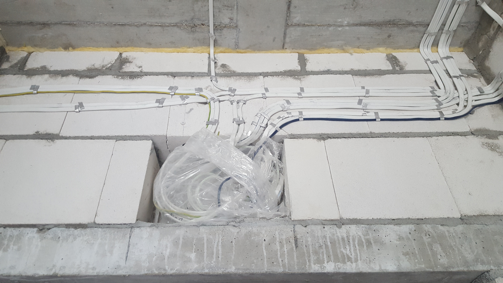 Białe przewody elektryczne prowadzone po ścianie z białych bloczków betonowych, zabezpieczone metalowymi uchwytami, część przewodów zwinięta w pętlę i zabezpieczona folią w otworze w ścianie.