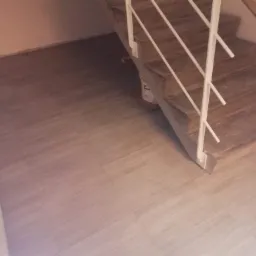 Klatka schodowa wykończona jasnymi panelami podłogowymi; widoczna biała balustrada i karton z klejem pod schodami.