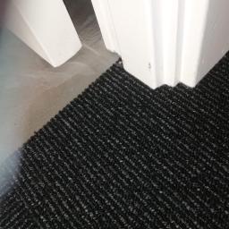 Jakub Majsterek - Fragment podłogi z czarną wykładziną dywanową przy białej listwie przypodłogowej, widoczny fragment betonowej wylewki.