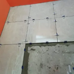 Układanie beżowych płytek podłogowych z użyciem systemu poziomowania, widoczna pomarańczowa ściana i surowy beton pod częścią płytek.