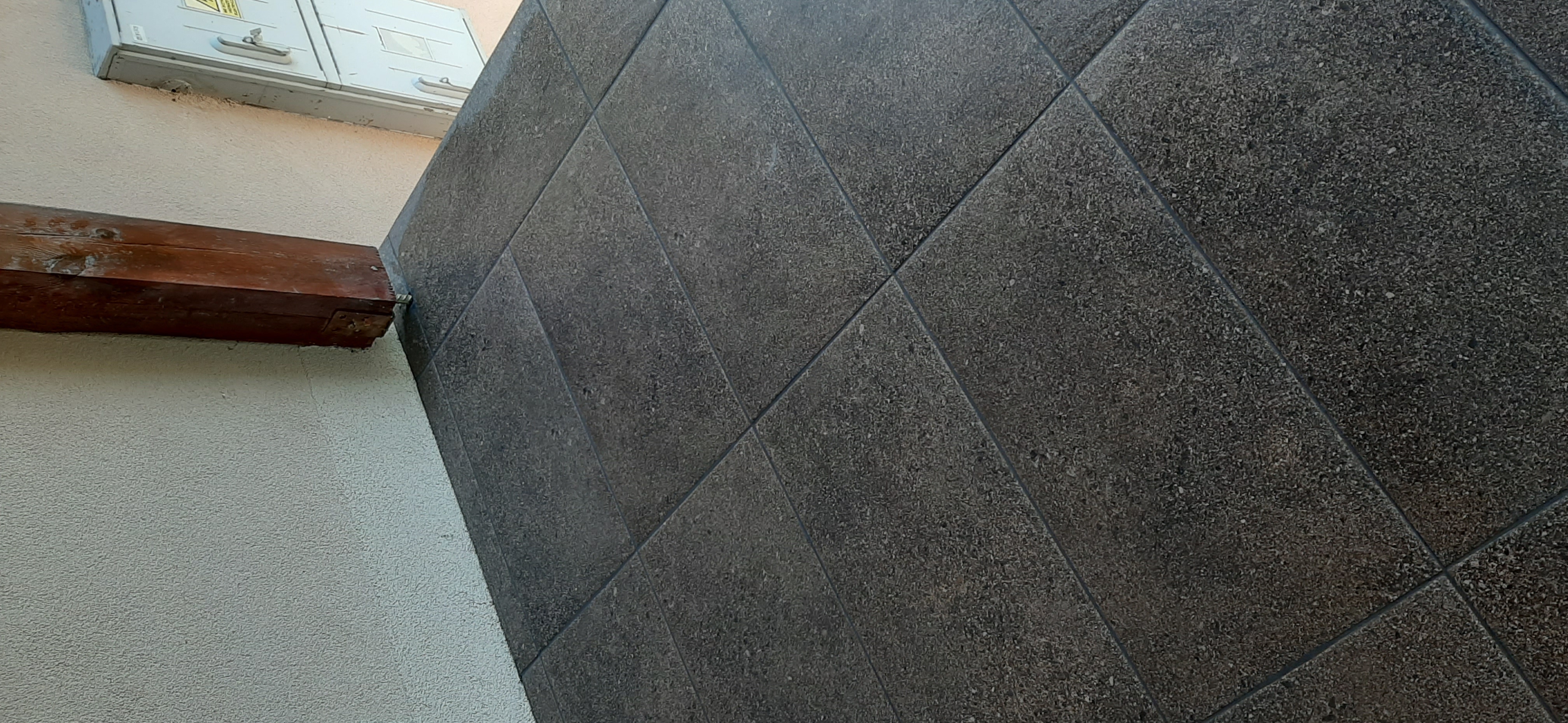 Ciemnoszare, kwadratowe płytki ułożone pod kątem na ścianie zewnętrznej budynku, widoczny fragment drewnianej belki i skrzynki elektrycznej.