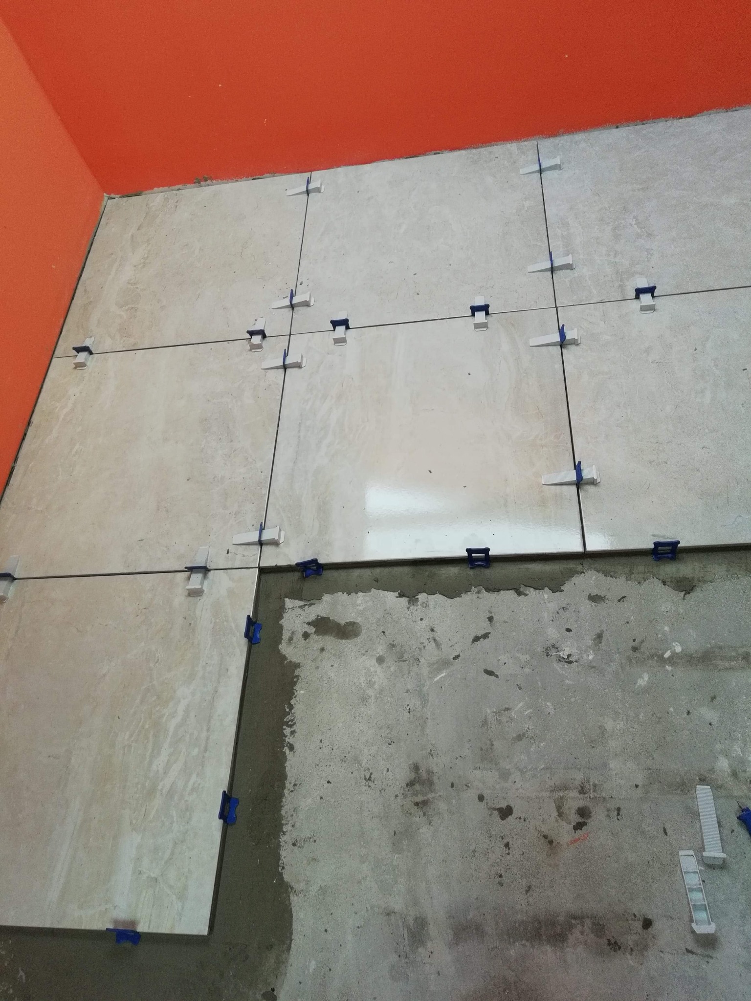 Układanie beżowych płytek podłogowych z użyciem systemu poziomowania, widoczna pomarańczowa ściana i surowy beton pod częścią płytek.