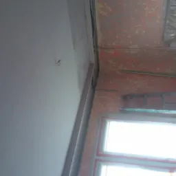 Fragment wnętrza w trakcie prac wykończeniowych: gładka, pomalowana na szaro ściana kontrastuje z nierówną, przygotowaną do dalszych prac ścianą w tle, widoczne okno i zabezpieczone folią elementy...