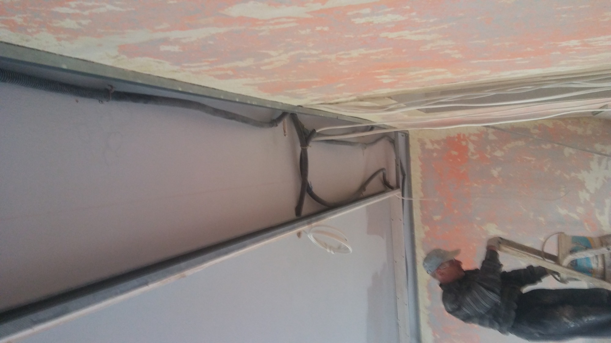 Instalacja sufitu podwieszanego: widoczne profile, kable w peszlach i pracownik na drabinie w trakcie montażu.