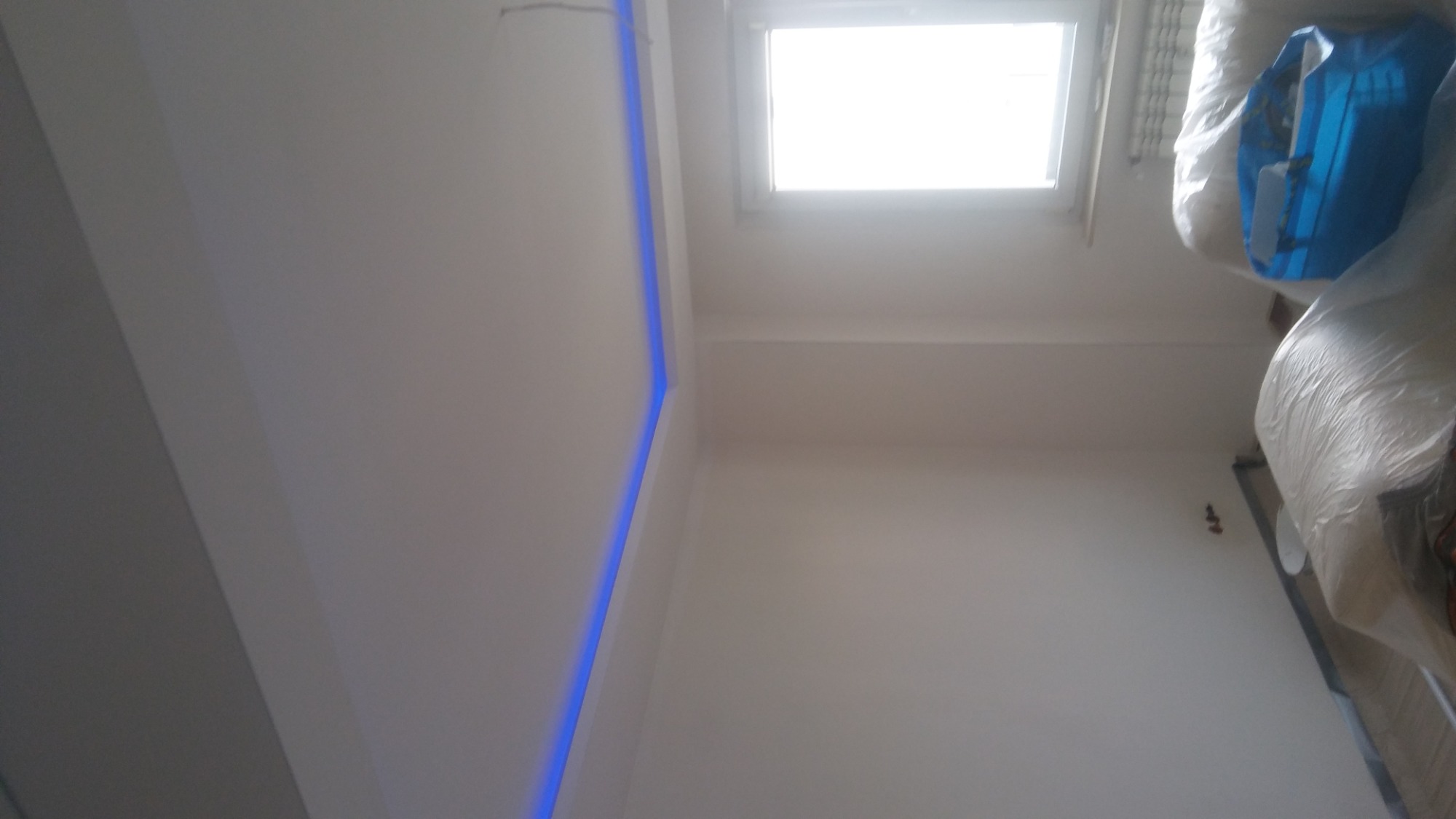 Ściana i sufit po szpachlowaniu z widoczną listwą LED w rogu, okno w tle, zabezpieczone meble folią malarską.