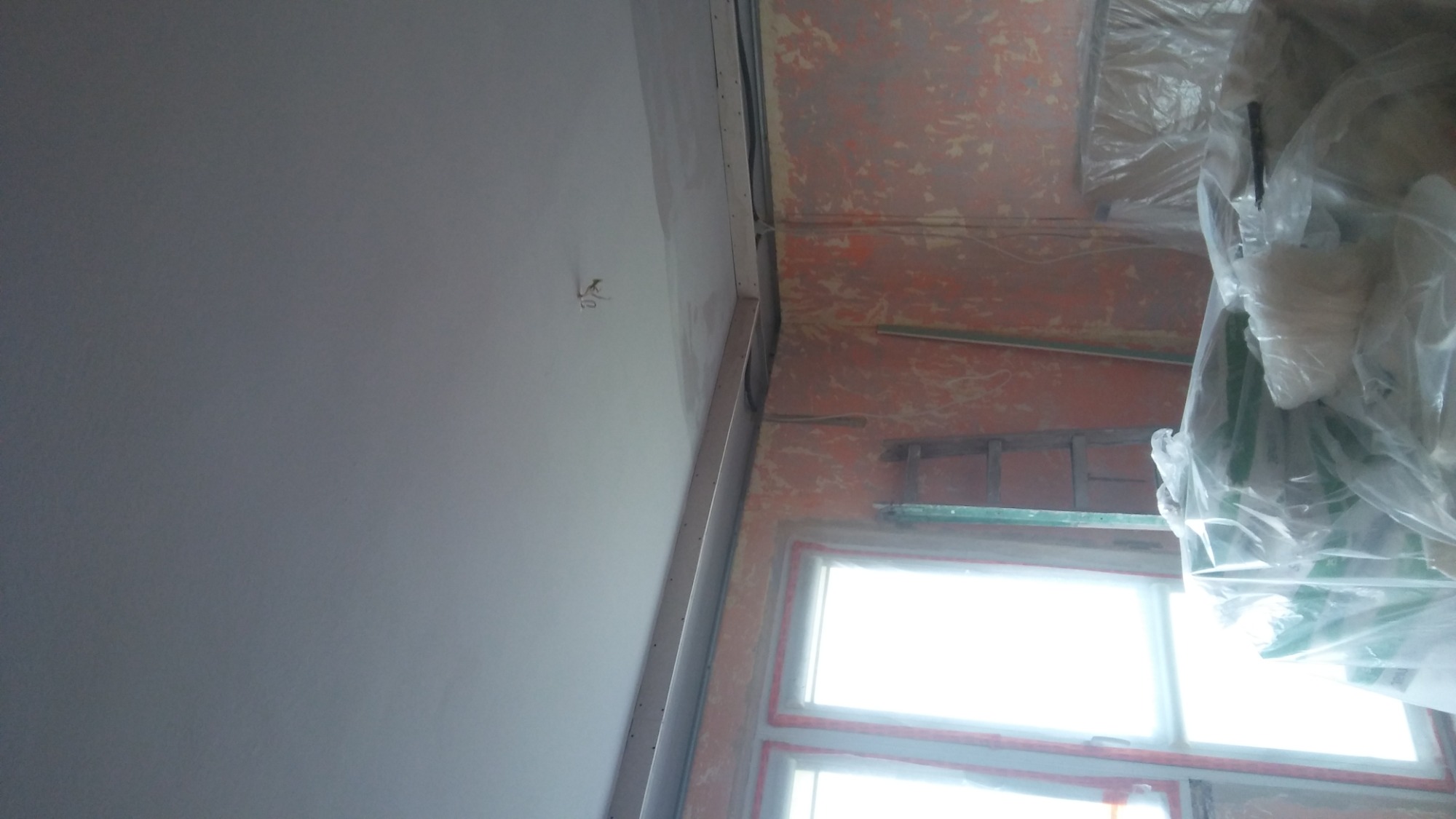 Fragment wnętrza w trakcie prac wykończeniowych: gładka, pomalowana na szaro ściana kontrastuje z nierówną, przygotowaną do dalszych prac ścianą w tle, widoczne okno i zabezpieczone folią elementy...