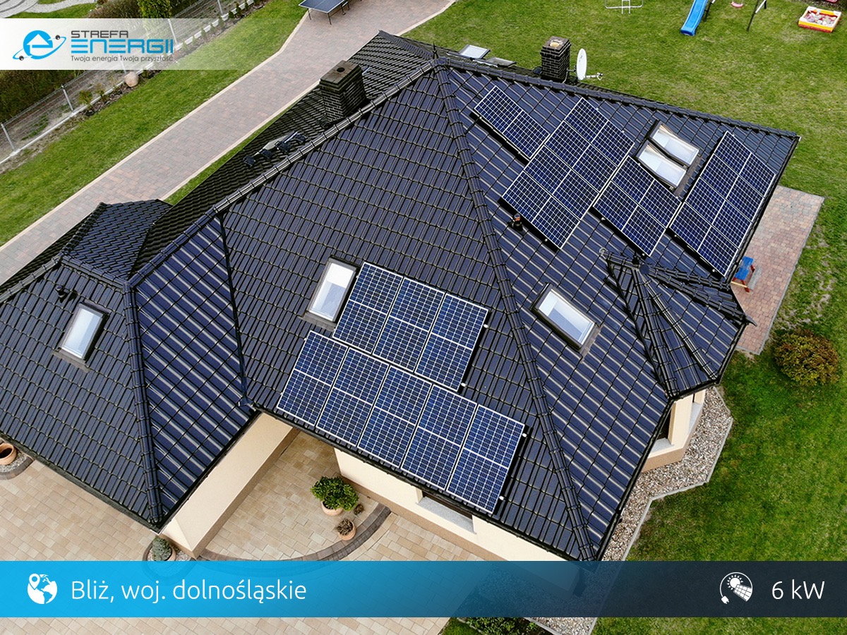 Widok z góry na dom z ciemną dachówką, na której zamontowano panele słoneczne i okna dachowe; trawnik i ścieżka z kostki brukowej wokół domu.