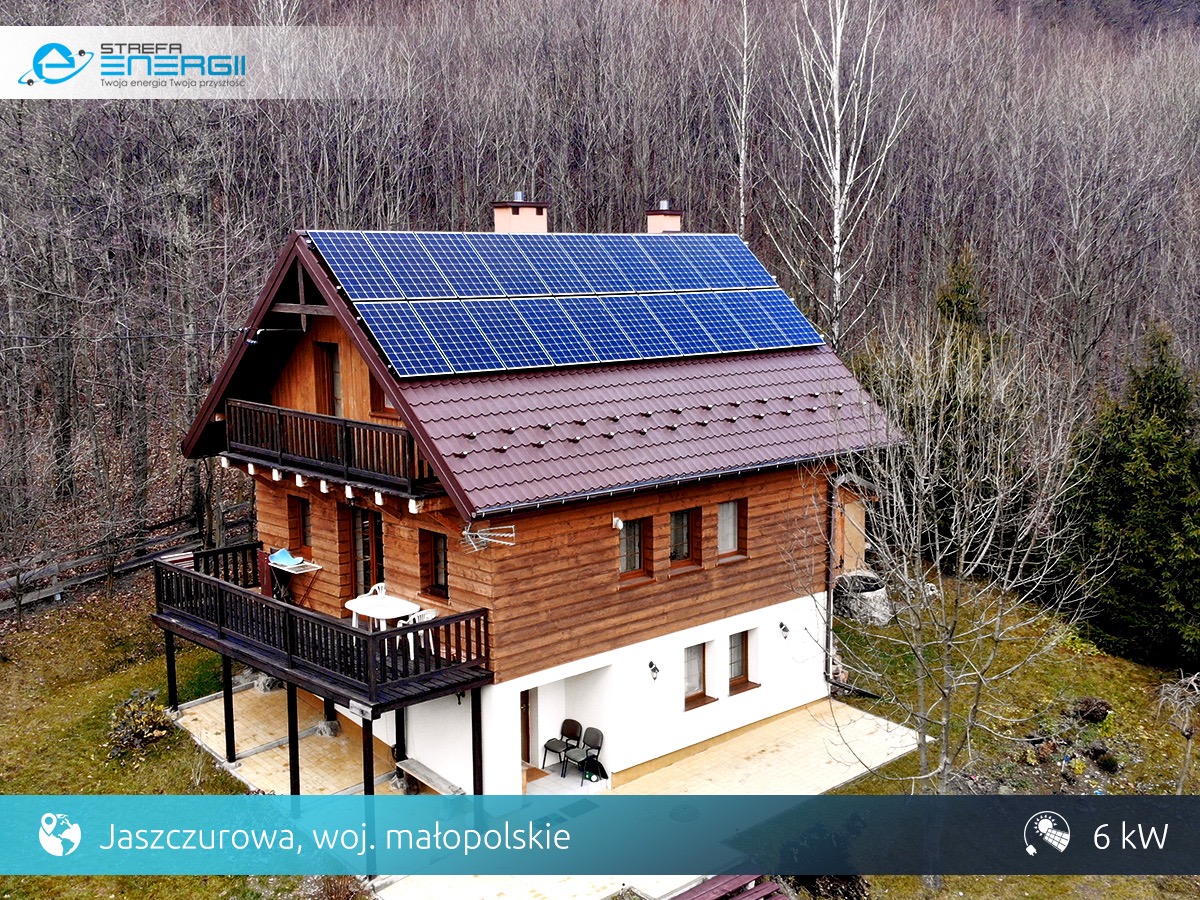 Dwukondygnacyjny dom z brązową dachówką i panelami słonecznymi na dachu, otoczony drzewami bez liści. Widoczny balkon z drewnianą balustradą i mały stolik.