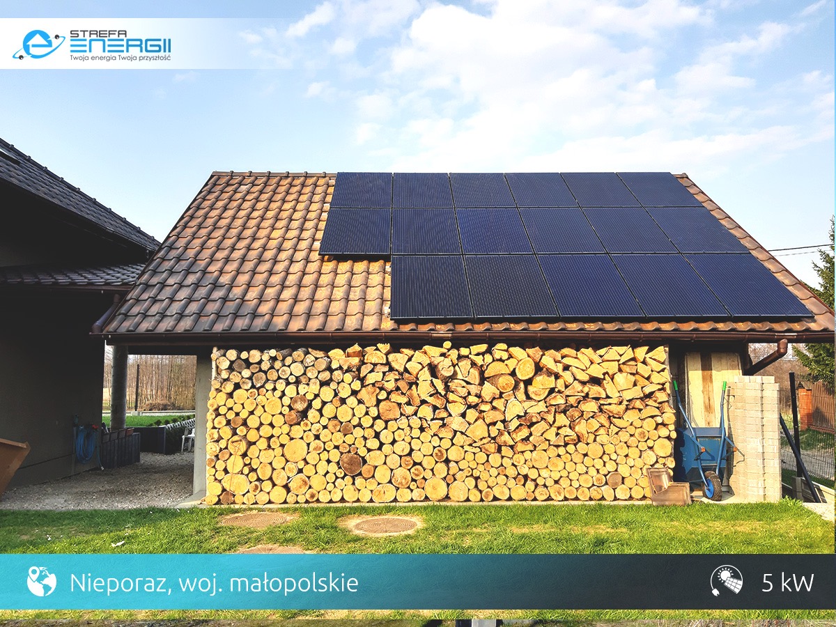 Instalacja fotowoltaiczna na dachu budynku z brązową dachówką, obok sterta drewna, w tle błękitne niebo z chmurami.