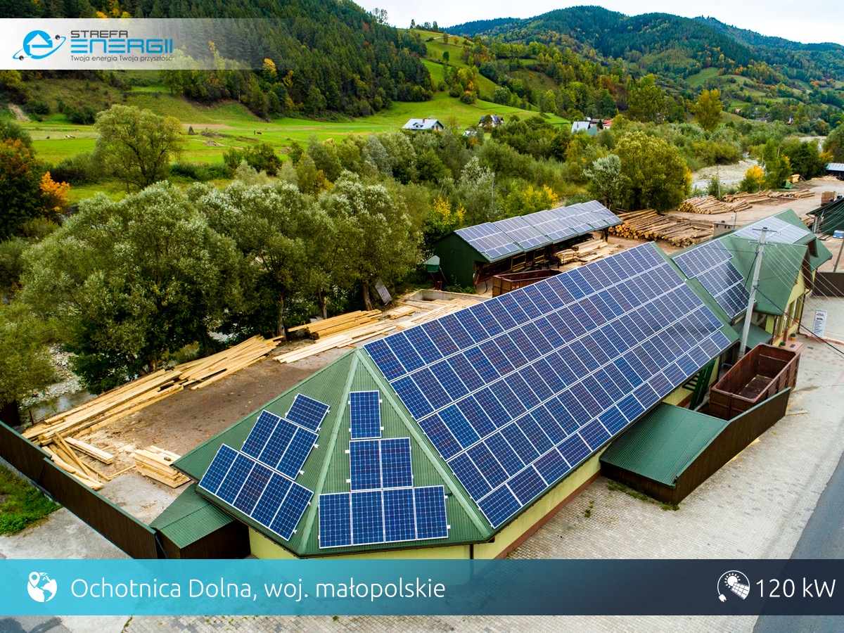 Widok z góry na budynek z zielonym dachem pokrytym panelami słonecznymi w Ochotnicy Dolnej, w tle górzysty krajobraz i składowane drewno.