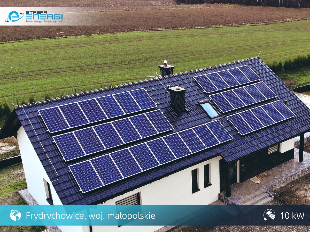 Dom jednorodzinny z czarnym dachem pokrytym panelami słonecznymi, widoczne okno dachowe i komin. W tle zielone pole.