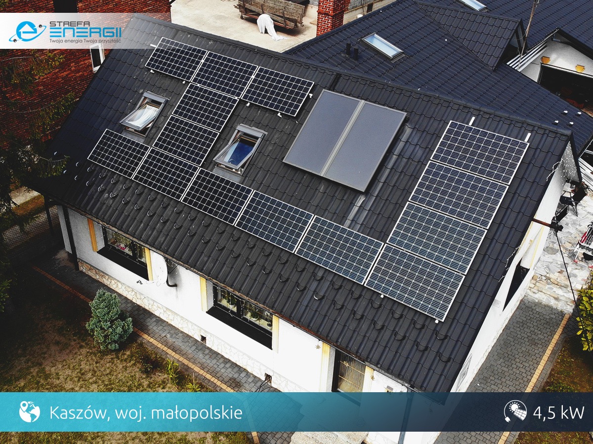 Widok z góry na dom z ciemnym dachem, na którym zamontowano panele słoneczne oraz kolektory słoneczne, widoczne okna dachowe.