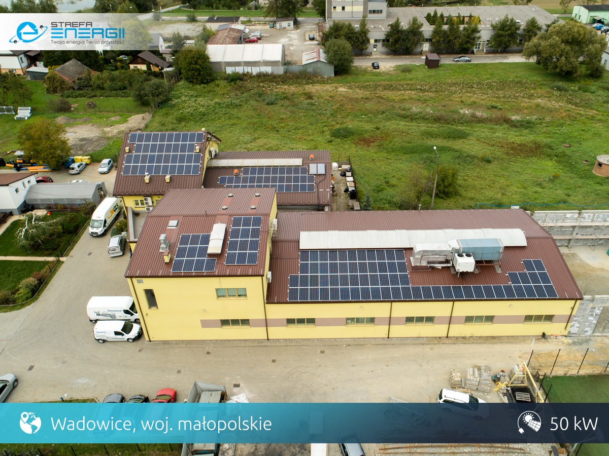 Widok z góry na budynki z panelami słonecznymi na dachach, teren firmy z zaparkowanymi samochodami dostawczymi, lokalizacja: Wadowice, woj. małopolskie, moc instalacji 50 kW.