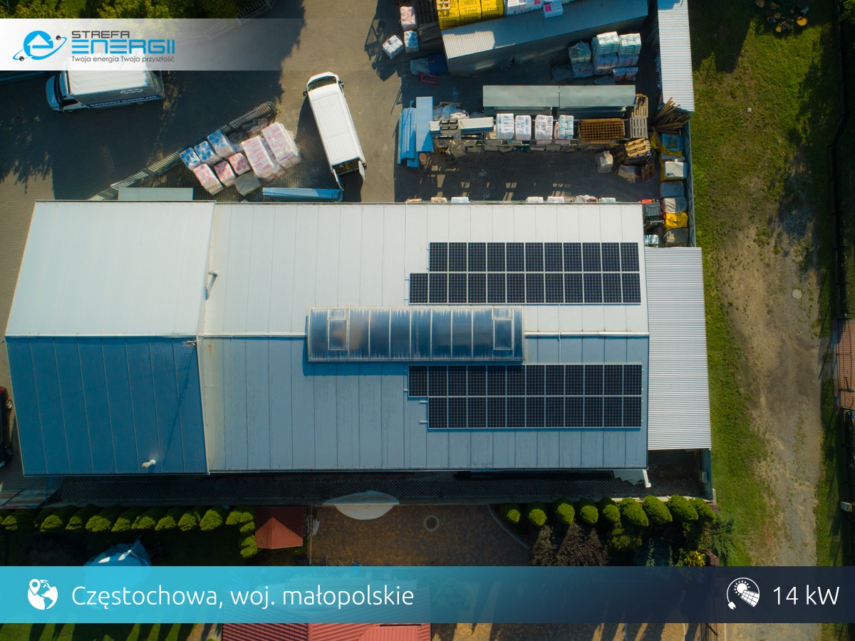 Widok z drona na halę przemysłową z zamontowanymi panelami słonecznymi, Częstochowa. Na dachu widoczne panele w dwóch sekcjach oraz świetlik. Obok hali zaparkowane dwa busy i składowane materiały...