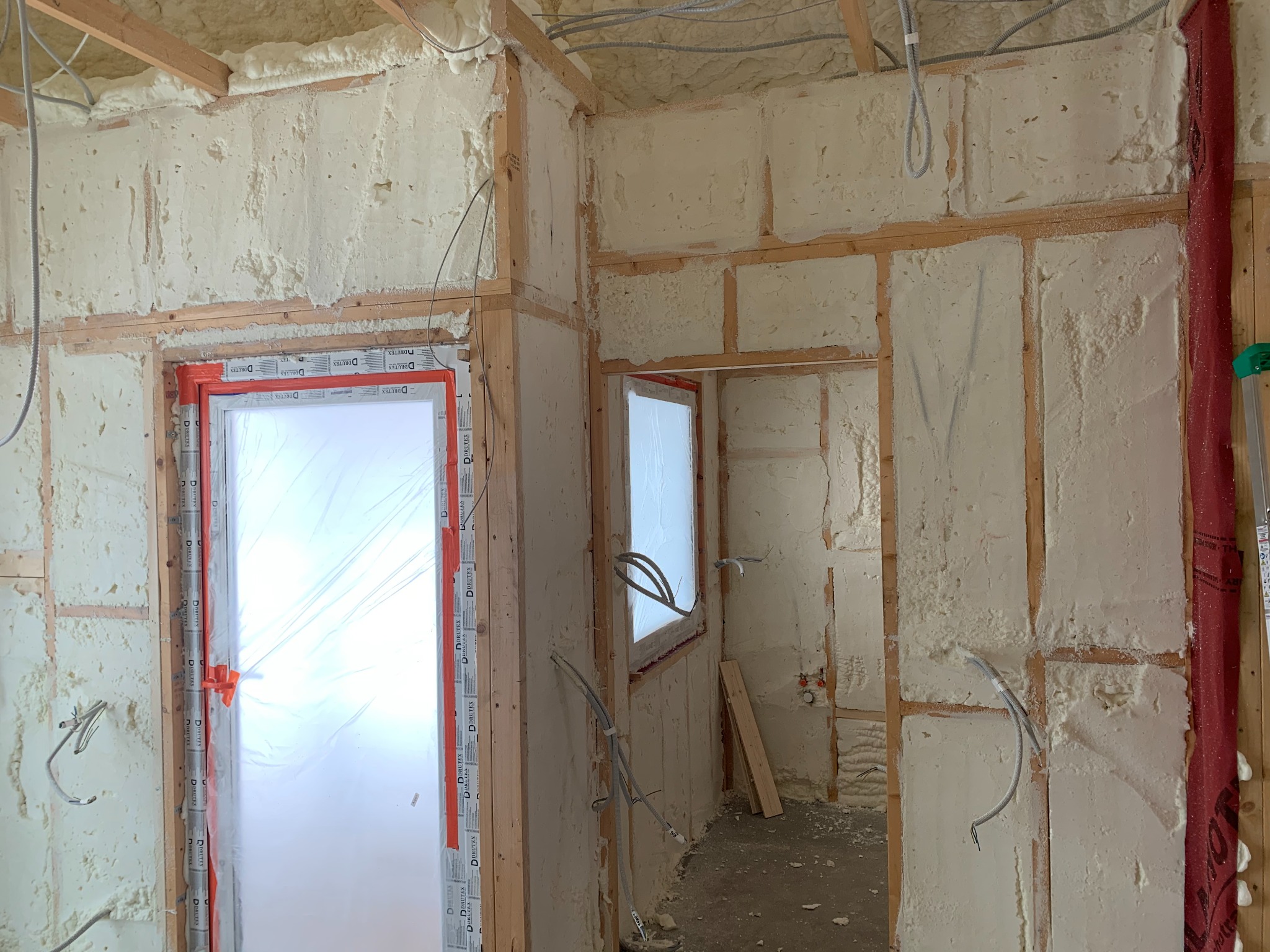 Wnętrze budynku w trakcie izolacji pianką PUR, widoczne drewniane ramy konstrukcyjne ścian wypełnione pianką, okno zabezpieczone folią ochronną, przewody elektryczne w peszlach.