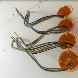 Cztery pomarańczowe puszki instalacyjne zamontowane w płycie kartonowo-gipsowej, z wystającymi przewodami o różnych kolorach, częściowo połączonych taśmą.