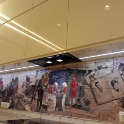 Maj Meble - Beżowa zabudowa kuchenna z podświetleniem LED i ozdobną ścianą nad blatem przedstawiającą kolaż zdjęć i grafik w stylu vintage.