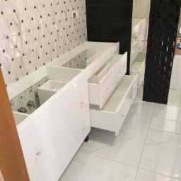 Biała szafka łazienkowa z otwartymi szufladami, kryształowe gałki, białe płytki podłogowe z szarymi żyłkami, dekoracyjne płytki ścienne w tle.