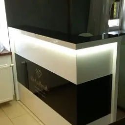 Nowoczesna lada recepcyjna w kolorze czarno-białym z podświetleniem LED, widoczne logo 'OrtoSmile' na czarnym panelu, jasne płytki podłogowe i fragment grzejnika w tle.