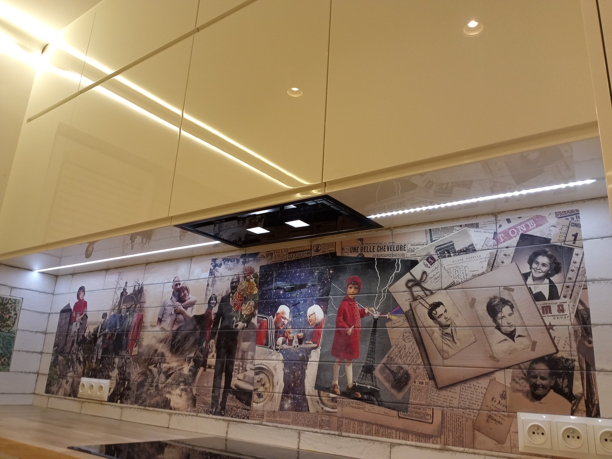 Beżowa zabudowa kuchenna z podświetleniem LED i ozdobną ścianą nad blatem przedstawiającą kolaż zdjęć i grafik w stylu vintage.