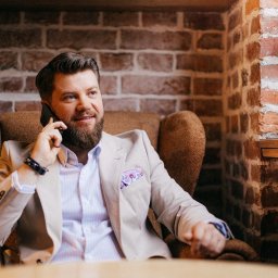 Ekspert Kredytowy Jakub Staszkiewicz - Mężczyzna w beżowym garniturze siedzi w fotelu na tle ceglanej ściany, rozmawiając przez telefon. Uśmiechnięty, z brodą, w jasnej koszuli i z poszetką w kieszonce.