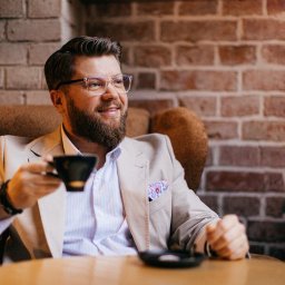 Ekspert Kredytowy Jakub Staszkiewicz - Uśmiechnięty doradca w beżowym garniturze, z filiżanką kawy na tle ceglanej ściany. Okulary z przezroczystymi oprawkami i wzorzysta poszetka dodają elegancji.