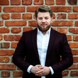 Portret mężczyzny z brodą w bordowej marynarce i białej koszuli na tle ceglanej ściany.