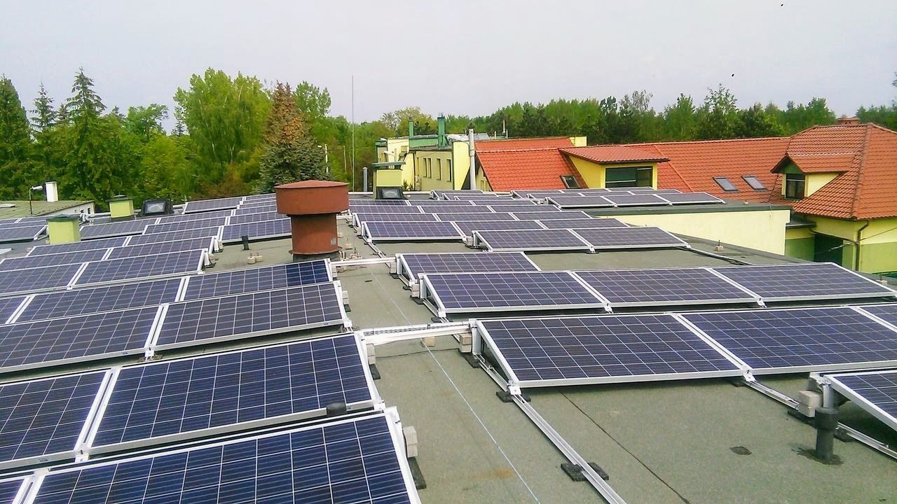 Widok z góry na dużą instalację paneli słonecznych na płaskim dachu budynku z zielonymi drzewami i budynkami w tle.