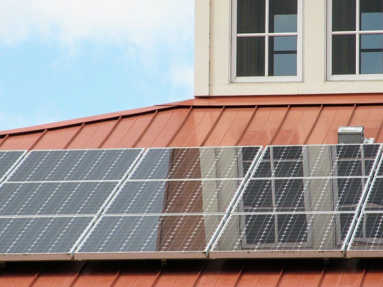 Panele słoneczne zamontowane na czerwonym blaszanym dachu domu z białymi oknami w tle, widoczne odbicie okien w panelach.