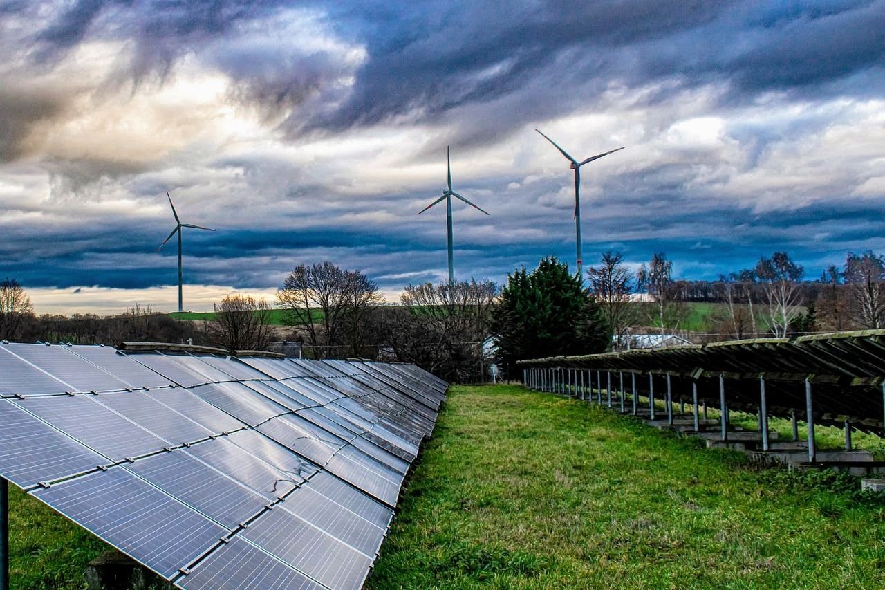 Rząd paneli słonecznych na tle pochmurnego nieba z turbinami wiatrowymi w oddali, widoczne odbicia w panelach.