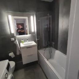 Nowoczesna łazienka z szarą glazurą, podświetlanym lustrem, białą szafką pod umywalką, narożną wanną z parawanem oraz pralką z przodu