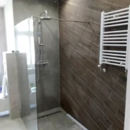 Nowoczesna łazienka z kabiną prysznicową typu walk-in, brązowymi płytkami ułożonymi w jodełkę na ścianie i białym grzejnikiem drabinkowym.