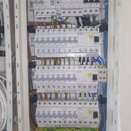 Instal Bart Bartłomiej Barylski - Rozdzielnica elektryczna z zainstalowanymi bezpiecznikami i okablowaniem w kolorach niebieskim, brązowym i zielono-żółtym, zamontowana w białej obudowie w ścianie w trakcie prac wykończeniowych.