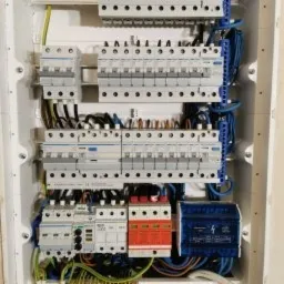 Rozdzielnica elektryczna w białej obudowie z widocznymi bezpiecznikami, wyłącznikami różnicowoprądowymi, listwami zaciskowymi i okablowaniem. Widoczne przewody w różnych kolorach. U góry i po...