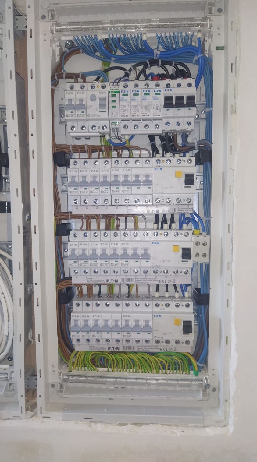 Rozdzielnica elektryczna z zainstalowanymi bezpiecznikami i okablowaniem w kolorach niebieskim, brązowym i zielono-żółtym, zamontowana w białej obudowie w ścianie w trakcie prac wykończeniowych.