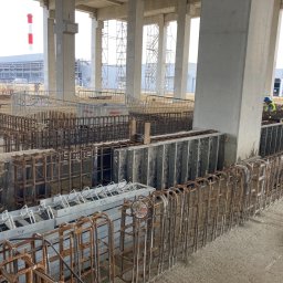 MMJ Construction Sp. z o.o. - Prace zbrojarskie w hali przemysłowej: widok na stalowe szkielety zbrojeniowe, przygotowane do zalania betonem, pracownicy w odblaskowych kamizelkach, betonowe filary hali w tle.