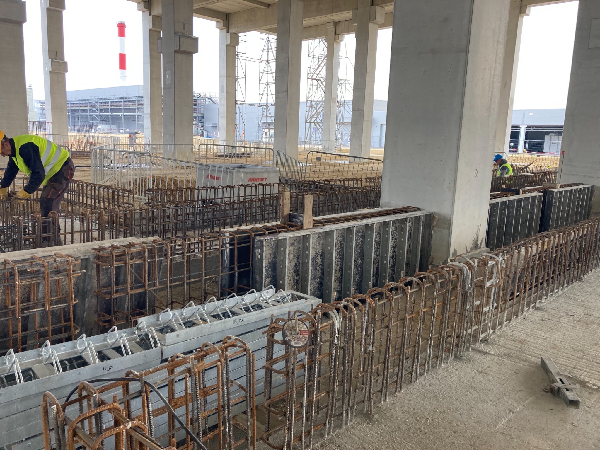 Prace zbrojarskie w hali przemysłowej: widok na stalowe szkielety zbrojeniowe, przygotowane do zalania betonem, pracownicy w odblaskowych kamizelkach, betonowe filary hali w tle.