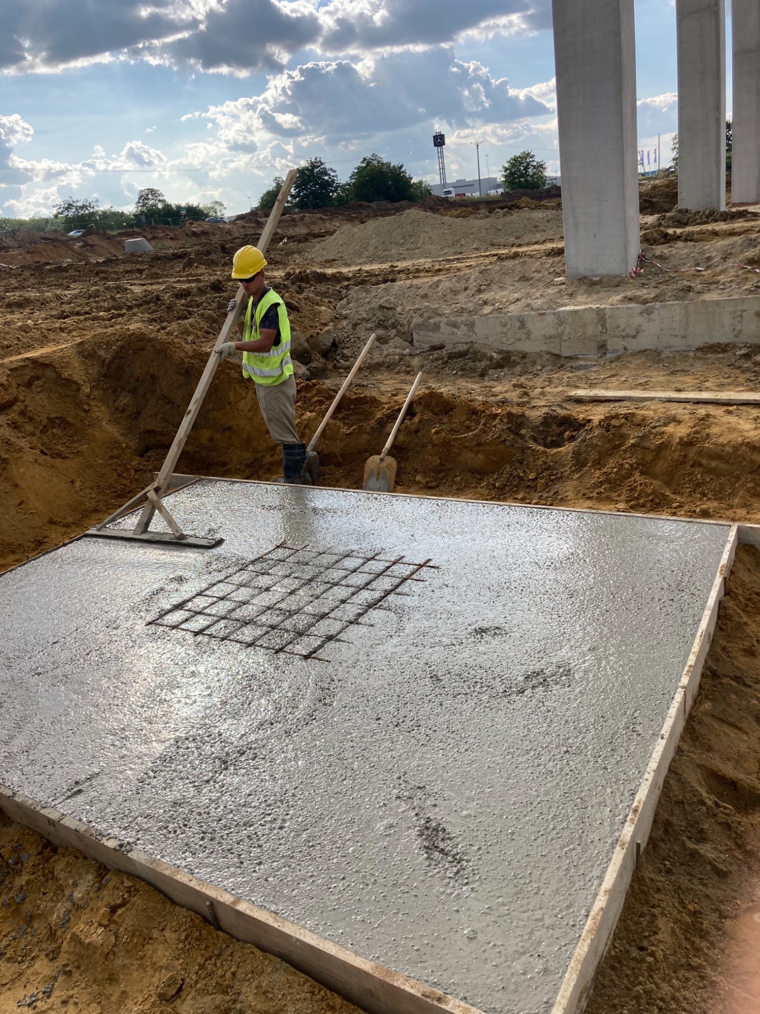 Pracownik w kasku i kamizelce odblaskowej rozprowadza świeży beton w szalunku za pomocą drewnianego narzędzia, widoczna siatka zbrojeniowa, w tle słupy konstrukcyjne i plac budowy.