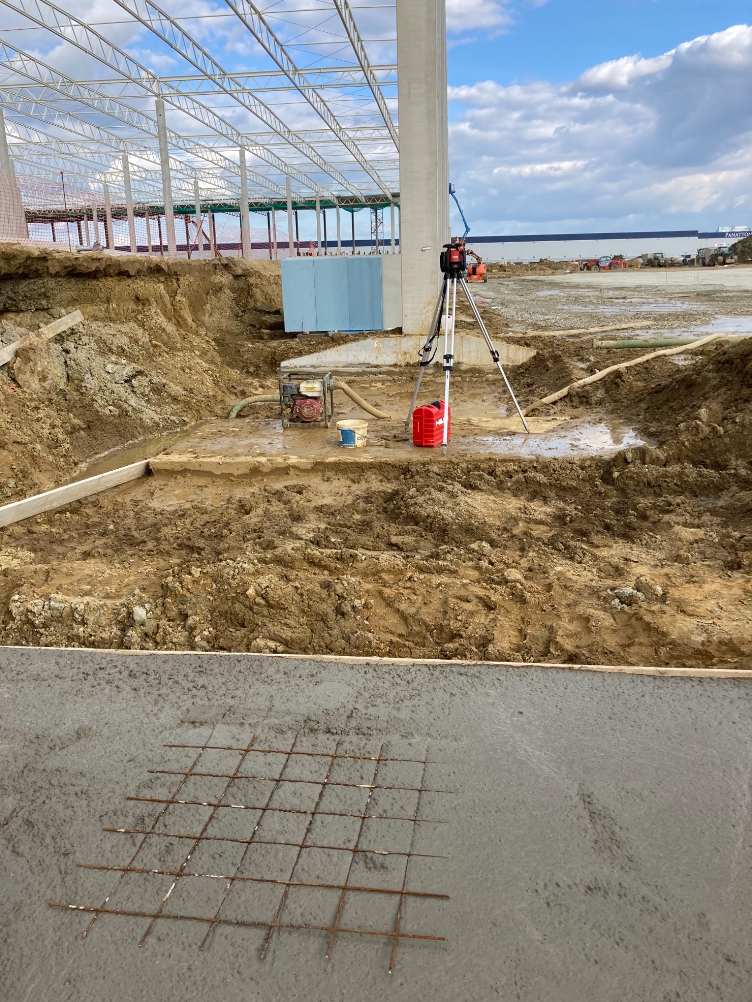 Wykop pod fundamenty hali stalowej, widoczny fragment zbrojenia w świeżym betonie, niwelator laserowy na statywie, pompa wodna i elementy izolacji w tle.